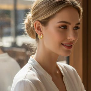 May include: A woman wearing gold hoop earrings. The earrings have a classic design with a smooth, polished surface. The woman is wearing a white blouse and has her hair styled in a bun. The background is blurred, suggesting natural light.
