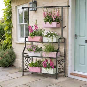 May include: A black metal tiered plant stand with scrollwork side details and a top hanging bar. The stand holds several rectangular wooden planter boxes in shades of pink, white, and green, filled with various blooming flowers and green foliage.