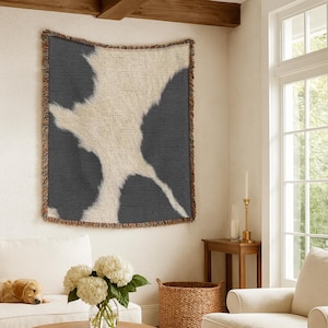 May include: A rectangular woven throw blanket featuring a high-contrast cowhide pattern in dark charcoal gray and off-white. The blanket has a textured fabric construction and is finished with decorative fringed edges along all four sides.