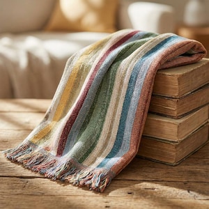 Puede incluir: Una colorida manta con rayas verticales en tonos amarillos, rojos, verdes, azules y marrones. La manta tiene un borde con flecos y está colocada sobre una pila de libros antiguos en una superficie de madera.