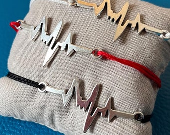 Könnte beinhalten: Drei silberfarbene Armbänder mit Herzschlag-Motiv und weißen, roten und schwarzen Bändern. Sie liegen auf einem kleinen, grauen Kissen vor blauem Hintergrund. Das Herzschlag-Design ist ein auffälliges Merkmal.