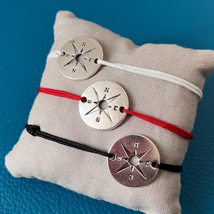 Peut inclure: Trois bracelets avec des breloques en forme de boussole argentées, avec des cordons blancs, rouges et noirs, sont présentés sur un coussin gris. Chaque breloque de boussole comporte des indications directionnelles.