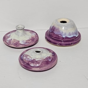 May include: Three handmade ceramic pieces in shades of purple and white. The pieces include two lids and a base, all with a glazed finish. The lids have a circular shape with a raised center, while the base has a rounded form with a series of small holes.