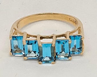 Puede incluir: Un anillo de oro con cinco piedras preciosas rectangulares de color azul claro. Las piedras están dispuestas en una fila en la parte superior del anillo. El anillo tiene un diseño sencillo y elegante.