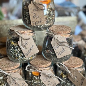 May include: A stack of small glass jars filled with dried herbs and spices, each sealed with a cork lid and tied with twine. The jars have small paper tags that read "Cold & Flu Relief", "Sleep + Relaxation", and "Menstrual Relief".