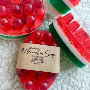 Könnte beinhalten: Handgemachte Wassermelonen-Seife in verschiedenen Formen. Die Seife ist rot mit schwarzen Kerndetails und einer grünen und weißen Schale. Eine Seife hat ein braunes Etikett mit der Aufschrift "Handmade Watermelon Soap".