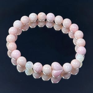 May include: A stretch bracelet composed of smooth, round, pale pink beads with a glossy finish. The bracelet features a single heart-shaped bead in the same soft pink hue, integrated into the circular design of the jewelry piece.