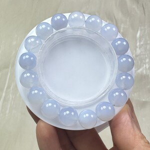 May include: A light blue beaded bracelet with round, polished beads. The bracelet is displayed on a white circular base with a clear plastic container in the center. The beads have a translucent quality.