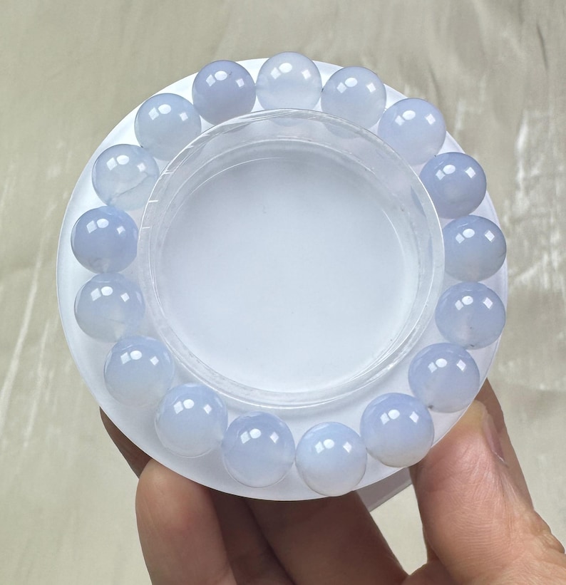 May include: A light blue beaded bracelet with round, polished beads. The bracelet is displayed on a white circular base with a clear plastic container in the center. The beads have a translucent quality.