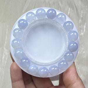 May include: A light blue beaded bracelet with round, polished beads. The bracelet is displayed on a white, circular container. The beads have a translucent quality, and the bracelet is held in a person's hand.