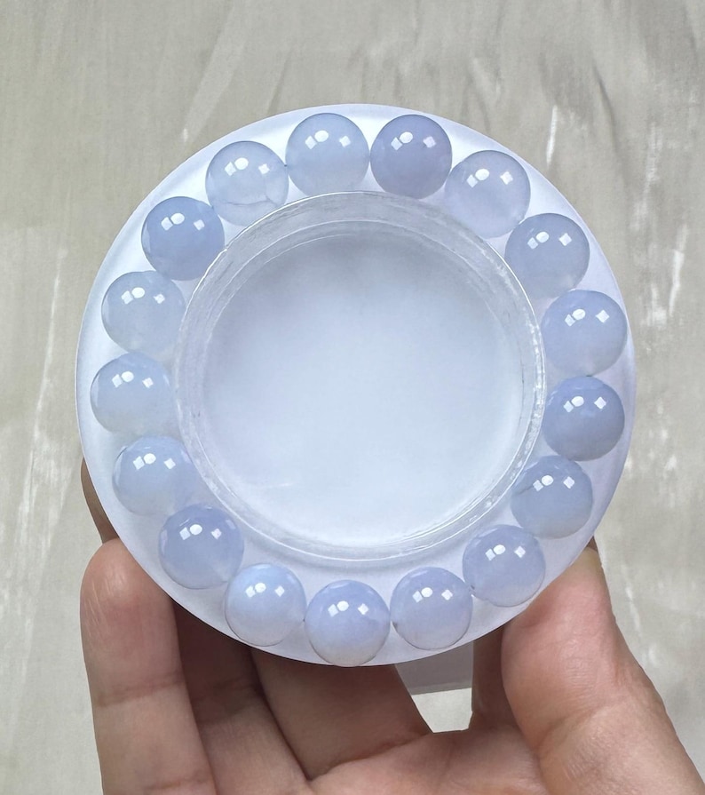 May include: A light blue beaded bracelet with round, polished beads. The bracelet is displayed on a white, circular container. The beads have a translucent quality, and the bracelet is held in a person's hand.