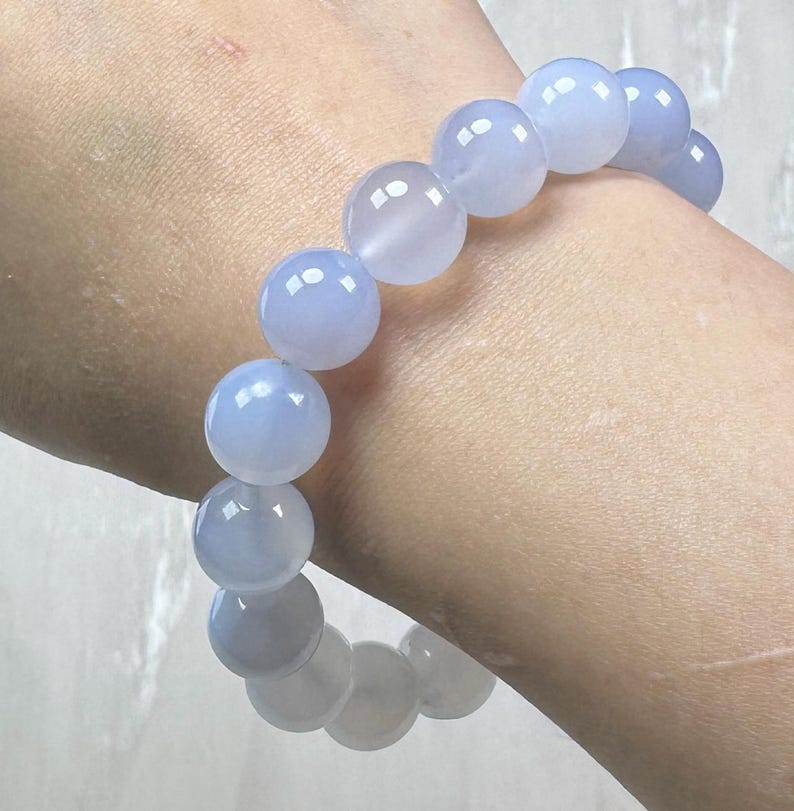 May include: A light blue beaded bracelet. The bracelet features large, round beads with a translucent appearance. The beads are strung together to form a complete circle. The bracelet is worn on a wrist.