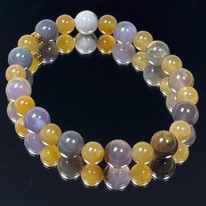 May include: A beaded bracelet featuring a mix of round beads in shades of yellow, purple, gray, and white. The bracelet has a gold-colored clasp and is displayed on a reflective black surface. The beads are approximately 0.5 inches in diameter.