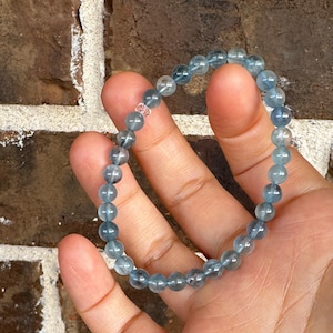 May include: A stretch bracelet composed of round, translucent blue beads with subtle internal inclusions. The beads are strung on an elastic cord, featuring a single clear, faceted spacer bead that interrupts the uniform pattern of the blue stones.