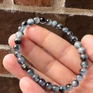 May include: A beaded bracelet featuring small, round, gray and blue beads. The bracelet is held in a hand, showcasing the natural variations in the stone's color and texture. The beads are strung together, forming a complete circle.