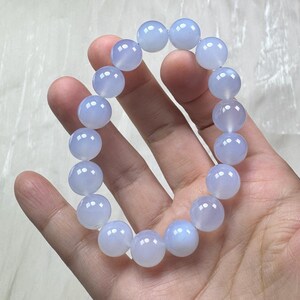 May include: A light blue beaded bracelet. The bracelet is made of round, translucent beads. The beads are a soft, pale blue color and appear to be polished. The bracelet is held in a hand against a white background. The bracelet is a piece of jewelry.