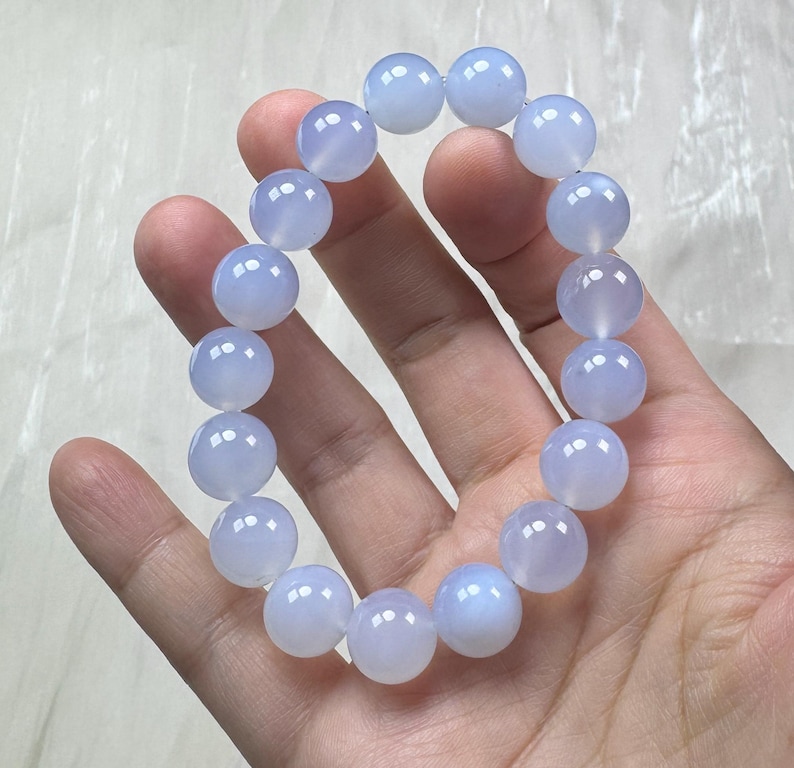 May include: A light blue beaded bracelet. The bracelet is made of round, translucent beads. The beads are a soft, pale blue color and appear to be polished. The bracelet is held in a hand against a white background. The bracelet is a piece of jewelry.