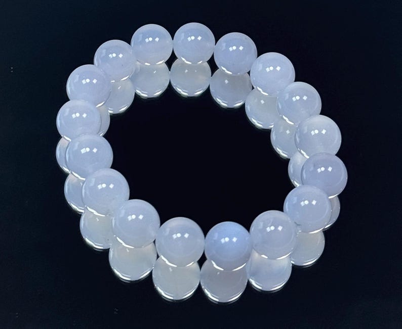 May include: A light blue beaded bracelet. The bracelet is made of round, polished beads. The beads have a translucent quality and reflect light. The bracelet is displayed on a reflective black surface.