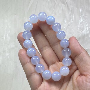May include: A light blue beaded bracelet. The bracelet is made of round, polished beads. The beads have a translucent quality and a soft, pale blue color. The bracelet is held in a hand against a white background.