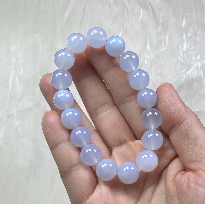 May include: A light blue beaded bracelet. The bracelet is made of round, polished beads. The beads have a translucent quality and a soft, pale blue color. The bracelet is held in a hand against a white background.