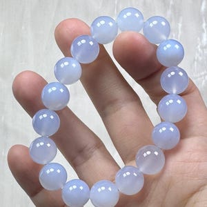 May include: A light blue beaded bracelet. The bracelet is composed of round, polished beads. The beads have a translucent quality, and the color is a soft, pale blue. The bracelet is held in a hand.