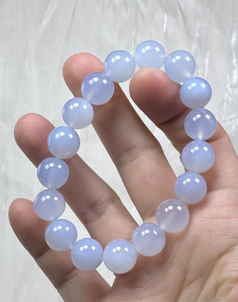 May include: A light blue beaded bracelet. The bracelet is composed of round, polished beads. The beads have a translucent quality, and the color is a soft, pale blue. The bracelet is held in a hand.