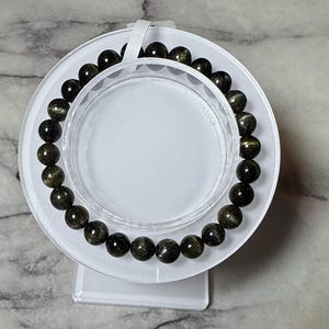 May include: A black and gold beaded bracelet with a shiny finish. The bracelet is displayed on a white stand.