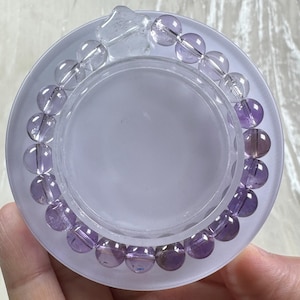 May include: A beaded bracelet featuring amethyst and clear quartz gemstones. The bracelet is composed of round, polished beads in shades of purple and clear, strung together on a clear elastic cord. The bracelet is displayed on a clear, circular stand.