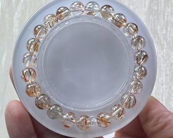Puede incluir: Una pulsera circular compuesta por cuentas de cuarzo rutilado pulidas y translúcidas. Cada cuenta presenta inclusiones distintivas en forma de aguja de color dorado y marrón oscuro suspendidas en la piedra clara, creando un patrón único.