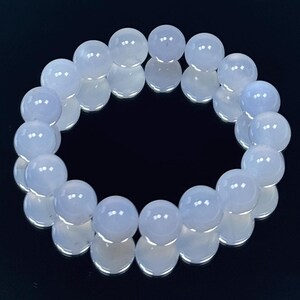 May include: A light blue beaded bracelet. The bracelet is made of round, polished beads. The beads are a soft, pale blue color and are strung together to form a circular shape. The bracelet is displayed on a reflective black surface.
