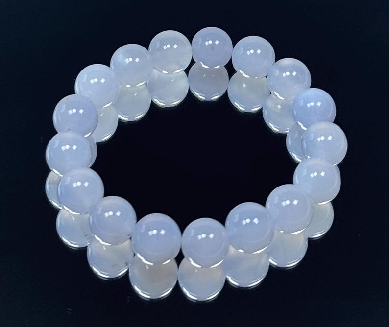 May include: A light blue beaded bracelet. The bracelet is made of round, polished beads. The beads are a soft, pale blue color and are strung together to form a circular shape. The bracelet is displayed on a reflective black surface.