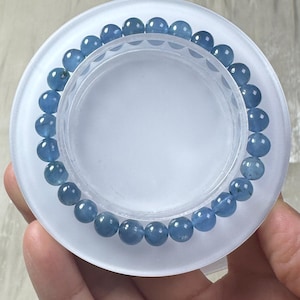 May include: A bracelet made of light blue, round beads. The beads are strung together on an elastic band. The bracelet is resting on a clear, circular display stand.