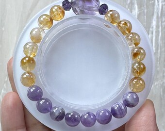 May include: A beaded bracelet featuring a mix of translucent golden yellow citrine spheres and smooth purple amethyst spheres. The bracelet is accented with a single large, faceted, irregular purple amethyst stone at the top, flanked by two small, dark purple faceted beads.