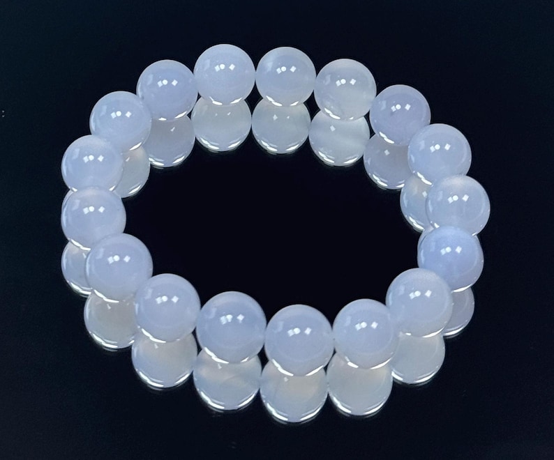 May include: A bracelet made of light blue, round gemstone beads. The beads are strung together to form a circular shape. The bracelet is displayed on a reflective black surface, enhancing the beads' color and shine.