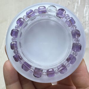 May include: A bracelet featuring amethyst beads in a variety of shapes and sizes. The bracelet includes faceted rectangular beads and smaller, round beads, all in shades of purple. The bracelet is displayed on a clear, frosted surface.