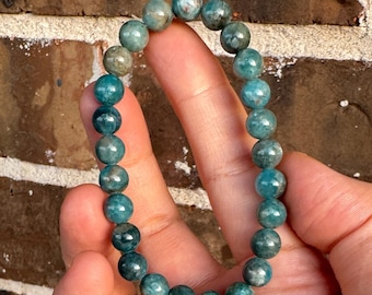 May include: A circular stretch bracelet composed of numerous smooth, round beads in varying shades of teal, blue-green, and dark forest green. The beads exhibit a polished, semi-translucent appearance with subtle internal marbling and natural variations.