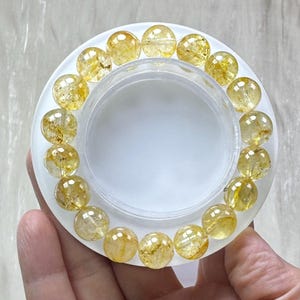 May include: A circular bracelet composed of translucent, golden-yellow rutilated quartz beads. Each spherical bead features internal needle-like inclusions and a polished, reflective surface, arranged in a continuous loop on a clear cord.