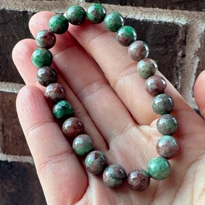 May include: A beaded bracelet featuring round, polished beads in shades of green and brown. The bracelet is strung together, showcasing the natural variations in color and texture of the stones. The bracelet is a piece of jewelry.