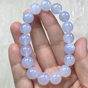 May include: A light blue beaded bracelet. The bracelet is made of round, polished beads strung together. The beads have a translucent quality, and the color is a soft, pale blue. The bracelet is held in a hand.