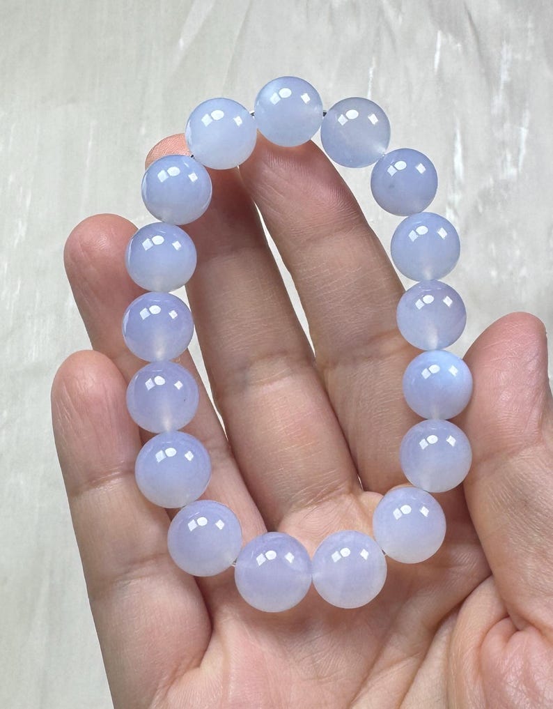 May include: A light blue beaded bracelet. The bracelet is made of round, polished beads strung together. The beads have a translucent quality, and the color is a soft, pale blue. The bracelet is held in a hand.
