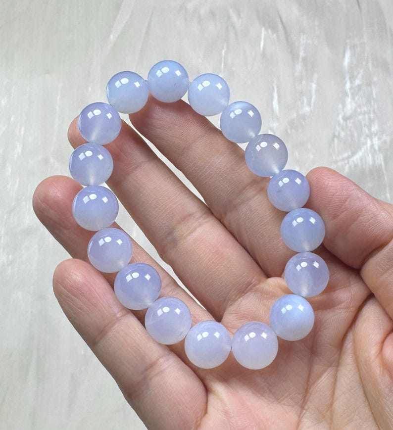 May include: A light blue beaded bracelet. The bracelet is made of round, polished beads. The beads are a soft, translucent blue color. The bracelet is held in a hand.