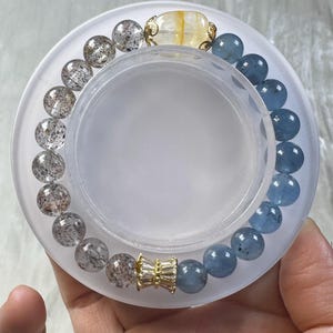May include: A beaded bracelet featuring a mix of clear, speckled, and blue-toned gemstones. The bracelet includes a gold-colored accent piece and a larger, yellow-toned stone. The bracelet is displayed on a white, circular surface.