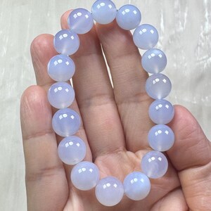May include: A light blue beaded bracelet. The bracelet is made of round, polished beads. The beads have a translucent quality and a soft, pastel blue color. The bracelet is held in a hand against a light background.