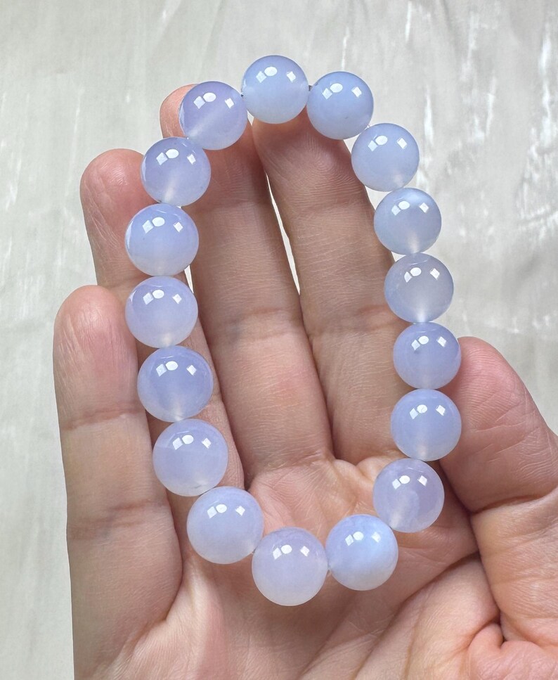 May include: A light blue beaded bracelet. The bracelet is made of round, polished beads. The beads have a translucent quality and a soft, pastel blue color. The bracelet is held in a hand against a light background.