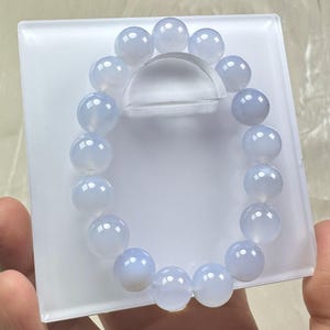 May include: A light blue beaded bracelet with large, round beads. The bracelet is displayed on a white, square platform with a clear, curved support. The beads have a translucent quality.