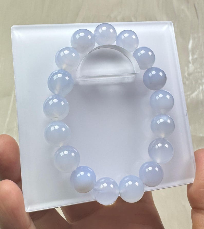 May include: A light blue beaded bracelet with large, round beads. The bracelet is displayed on a white, square platform with a clear, curved support. The beads have a translucent quality.