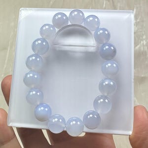 May include: A light blue beaded bracelet. The bracelet is composed of round, polished beads. The beads are a soft, translucent blue color. The bracelet is displayed on a white, rectangular surface.