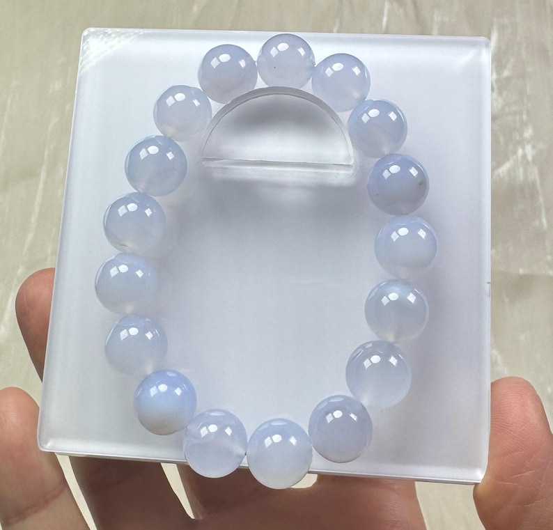 May include: A light blue beaded bracelet. The bracelet is composed of round, polished beads. The beads are a soft, translucent blue color. The bracelet is displayed on a white, rectangular surface.