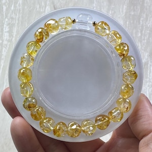 May include: A circular bracelet composed of faceted, translucent yellow citrine beads. The beads feature internal golden inclusions and are strung on a clear elastic cord, secured with a small gold-toned metal bead near the clasp area.