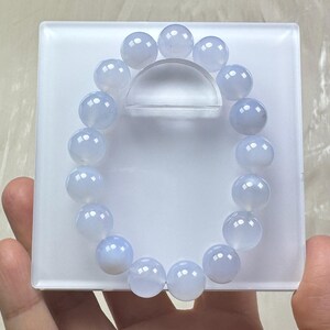 May include: A light blue beaded bracelet with round, polished beads. The bracelet is displayed on a white, rectangular stand with a clear, curved support. The beads have a translucent quality, and the bracelet is held by a hand.